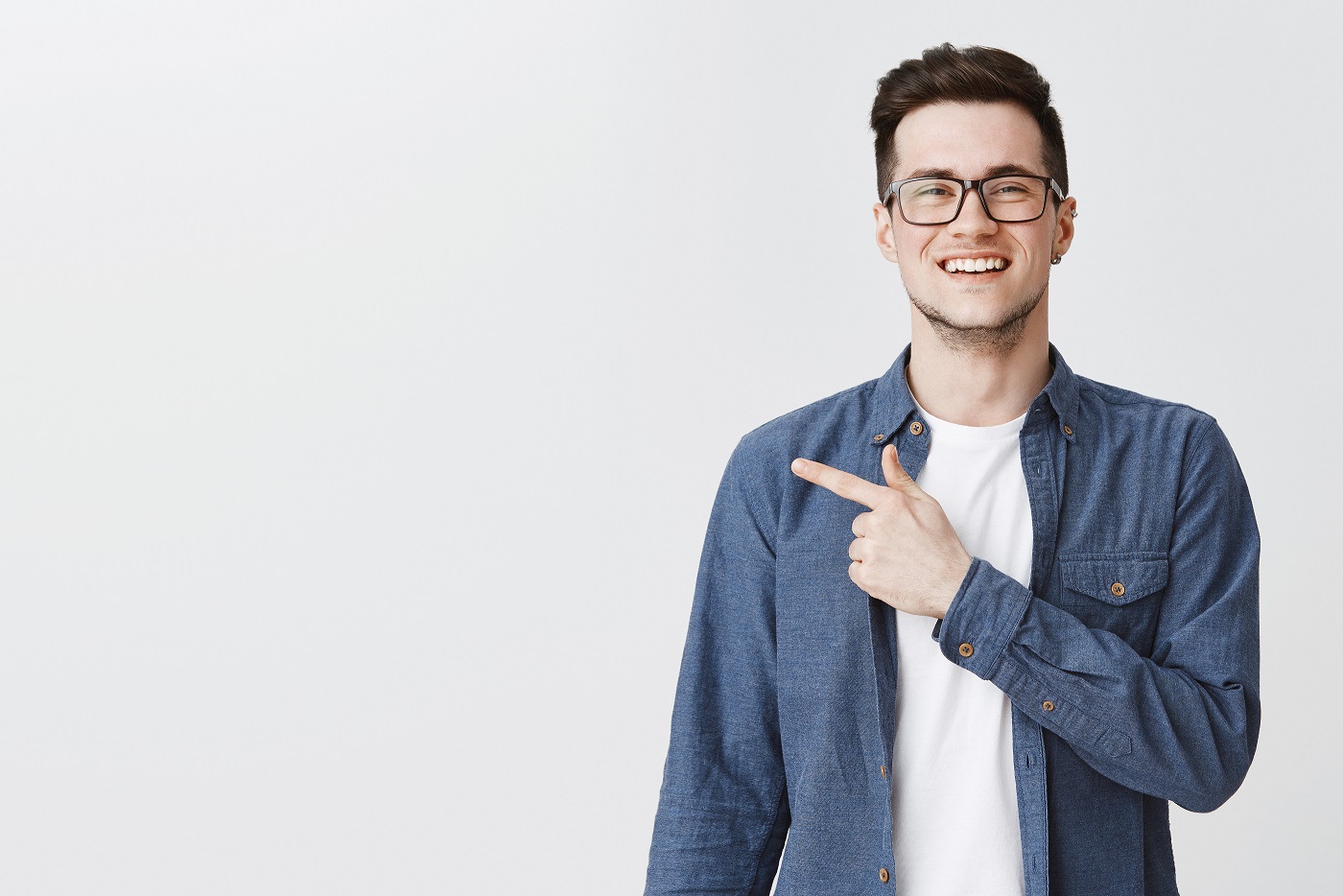 Polite and friendly handsome young caucasian man in glasses and blue shirt over t-shirt smiling joyfully as pointing left at copy space showing cool place for advertisement over grey wall. Emotions concept